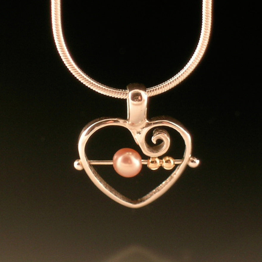 Close up view of pendant on Abacus Heart Pendant. This pendant is in the shape of a heart with a swirl inside the top right corner. There is a silver bar running through the center of the heart (horizontally) with two small beads of gold on the right and a bigger pink pearl bead on the left.