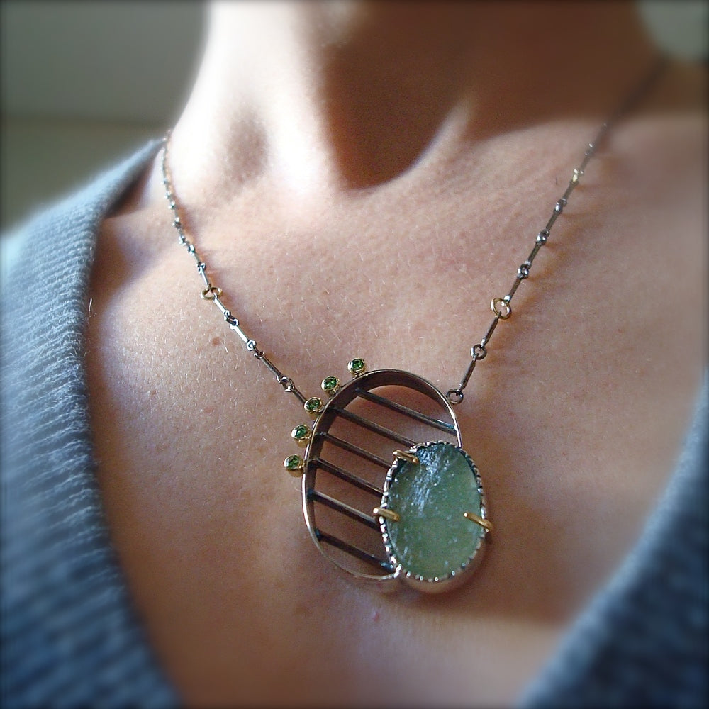 Full view of Striated Necklace - green beryl and Green Diamonds on woman's neck to give idea of scale of piece. the silver pendant lays on a hand fabricated silver chain. The pendant is a silver oval with silver lines running through it. The bottom right corner of the oval has a set piece of raw green beryl and on the opposite side are five set green diamonds lining the silver oval.