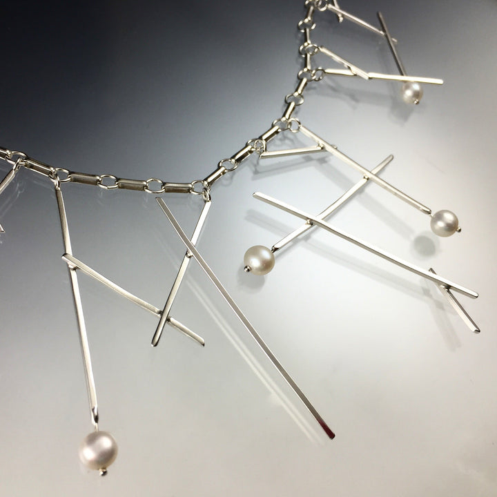 Close up of neckline of Sticks and pearls Necklace. this necklace is made of silver and has silver square wire hanging from its chain resembling sticks and beaded pearls on some of the ends of the "sticks".