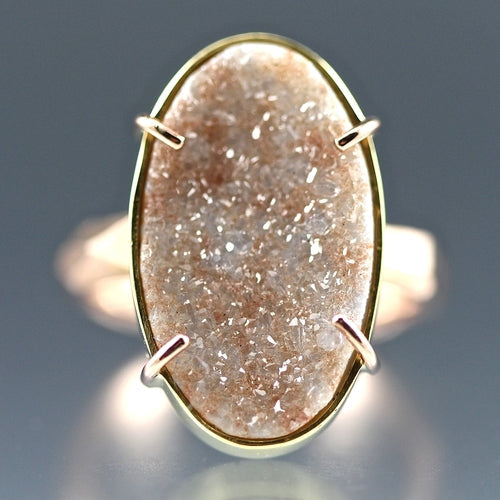 Detail shot of gem on Peach Druzy Chiseled Ring. This peach druzy gem is cut in the shape of an oval, set with four rose gold prongs on a gold bezel on a rose gold band.