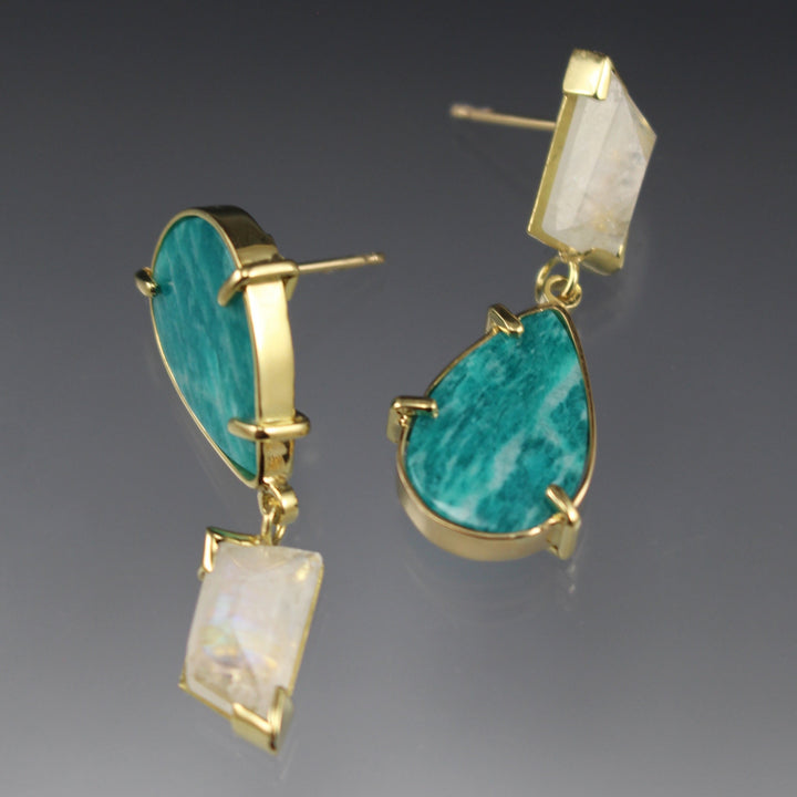 Full image of angled view Amazonite and Moonstone Asymmetrical Earrings. These earrings have the amazonite in the shape of a teardrop and the moonstone in an asymmetrical rectangle all encompassed in gold. These earrings are asymmetrical in that the moonstone and amazonite are flipped in the corresponding earring.