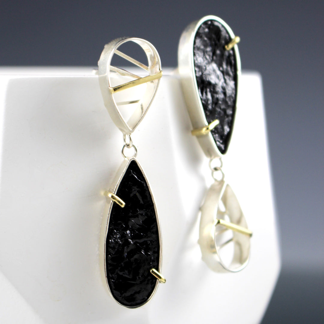 Angled view of Asymmetrical Earring with Black Tourmaline. These earrings are in the shapes of teardrops, a smaller on made of a silver border and silver and one gold bar crossing through the outline. The secondary teardrop is of the raw black tourmaline that is encompassed in silver with two gold opposing prongs. The asymmetrical look comes from the teardrops opposing each other compared to its matching earring.