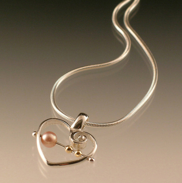 Angled view of Abacus Heart Pendant. This pendant is in the shape of a heart with a swirl inside the top right corner. There is a silver bar running through the center of the heart (horizontally) with two small beads of gold on the right and a bigger pink pearl bead on the left.