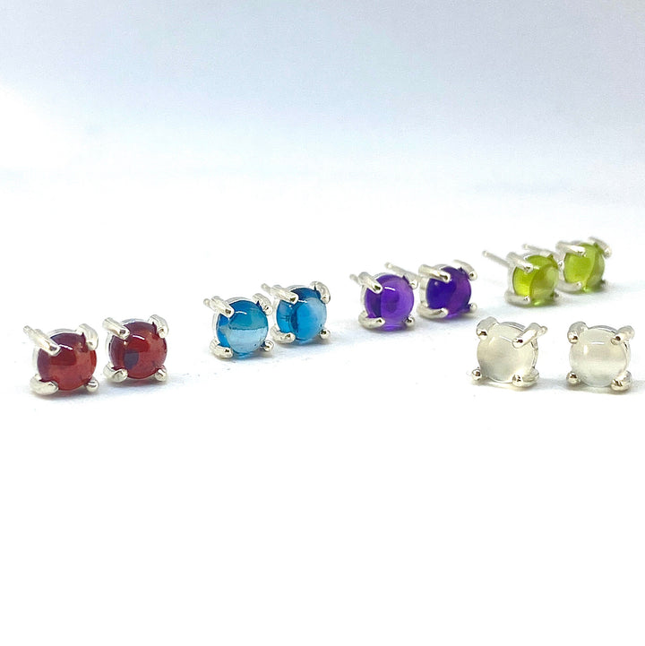 Full view of five different colored (maroon, light blue, navy blue, yellow-green, and clear) small round stud cabochon earrings.