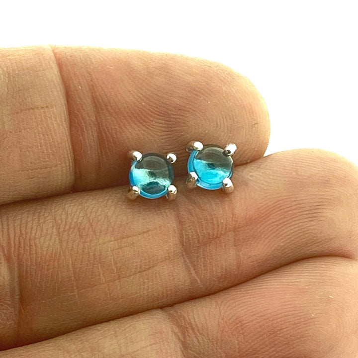 Full view of light blue small round cabochon stud earrings in-between two fingers to help give idea of scale of piece.