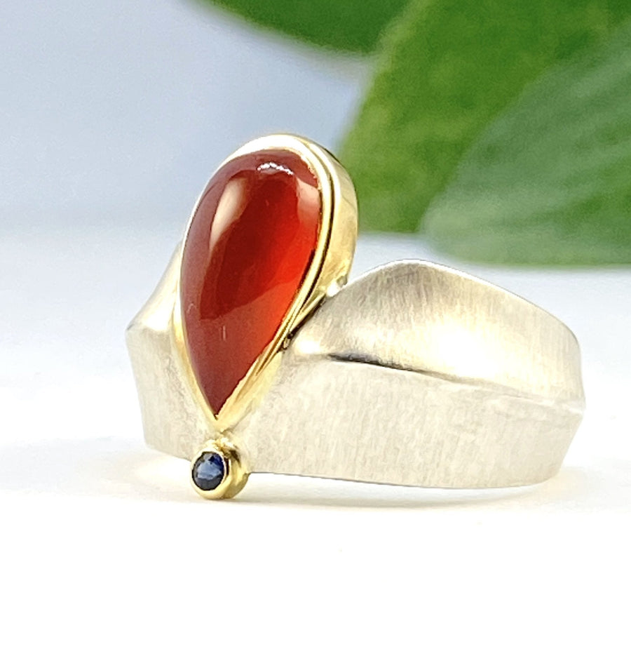 Angled view of Carnelian and Sapphire Ridge Ring. This ring showcases a rain drop shaped carnelian encompassed in gold on a fat silver band. Across the carnelian is a set blue sapphire in gold and right at the tip of the teardrop.